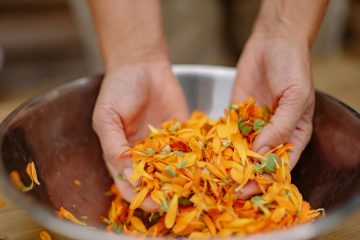 Close-up image of hands holding vibrant orange flower petals in a steel bowl, showcasing nature's colors.