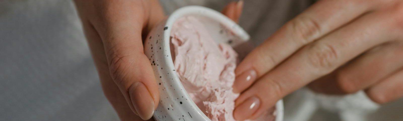 Close-up of hand applying pink moisturizing cream from a ceramic bowl for skincare routine.